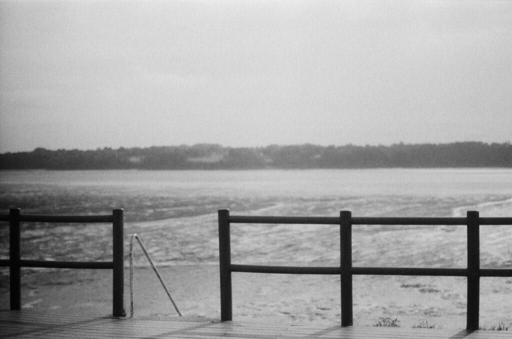 Black and white film photograph of an open fence leading to a body of water.